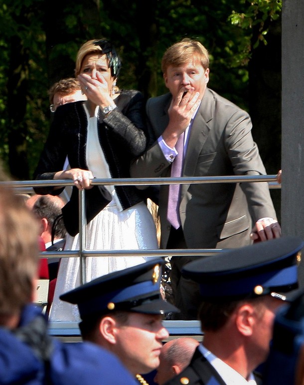 Photo 2 Apeldoorn 30 of April 2009: Maxima clamps her one hand on the railing of the bus and the other over her mouth and nostrils, as she watches the totally unexpected terrible scene in front of her; Willem Alexander is captured with his mouth visible through his fingers in an inelegant way, one of the rare moments were his trained  official mask has fallen of; their eyes survey the disastrous scenery but also seem to be turned inward, thoughts racing through their minds; "was this aimed at us?"; seeing the victims on the street and people rushing up to help them; seeing the crashed car; curious is that the face of the one policeman that can be seen in full profile looks the other way, as if he has not seen what has just happened and it is a face without any sign of having witnessed a disaster; it could be that as the policemen stand lower their view is blocked; nobody is seen in a pose of shrinking back from an approaching danger, or pulling themselves together after such a fear. The overall impression is that of spectators in awe.